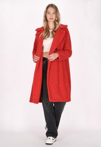 Cappotto teddy rosso e morbido con colletto a fazzoletto, chiusura con un solo bottone e taglio A-line, abbinato a jeans neri a vita alta e sneakers bianche.