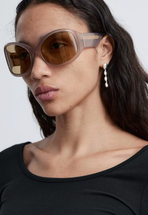 Vrouw met lang donker haar, oversized bruine zonnebril, zilveren druppelvormige oorbellen en een zwart topje met een brede halslijn.