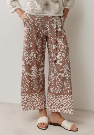 Wide-leg pants with brown and white tropical and animal print, paired with cream knit sweater and white slip-on sandals. Hands in pockets.