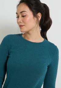 Blusa de manga larga color verde azulado con tela texturada y cuello redondo. Presenta patrones sutiles y es ajustada en la cintura.