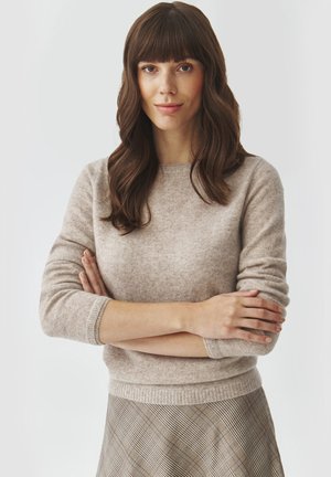 Beige Strickpullover mit Rundhalsausschnitt und langen Ärmeln, kombiniert mit einem karierten Rock in Creme-, Braun- und Grautönen.