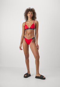 Rød bikini sett med trekantet bralette og matchende lave bunner, med en svart logo-stripe. Glatt, skinnende tekstur.