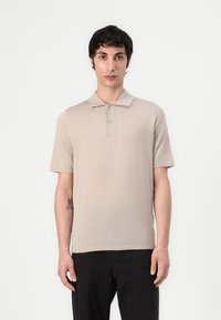 Polo beige à manches courtes avec un col plat, doté d'une patte de boutonnage à trois boutons et d'un tissu fin et texturé. Porté avec un pantalon sombre.