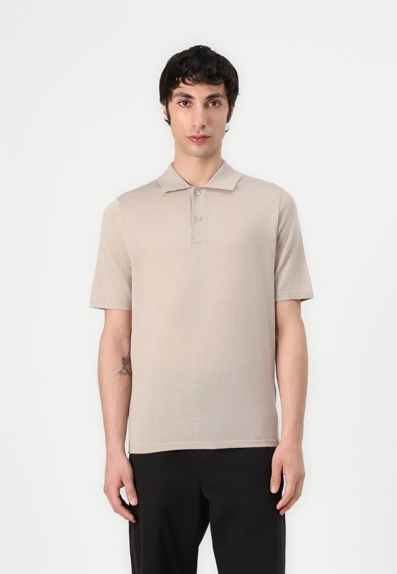 Polo beige à manches courtes avec un col plat, doté d'une patte de boutonnage à trois boutons et d'un tissu fin et texturé. Porté avec un pantalon sombre.