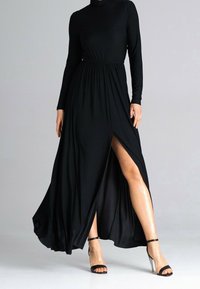 Zwarte maxi-jurk met lange mouwen, hoge hals en een zijsplit. Gemaakt van soepel stof met een elastische taille en een vloeiende rok. Gecombineerd met zwarte sandalen met bandjes.