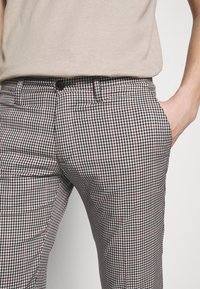 Personne portant un pantalon à carreaux noir, blanc et rouge avec une chemise beige rentrée, main droite dans la poche.