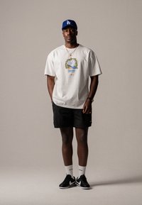 T-shirt en coton blanc avec un design graphique coloré, shorts noirs et baskets noires avec des accents blancs. Casquette bleue et chaussettes blanches arrivant aux chevilles.