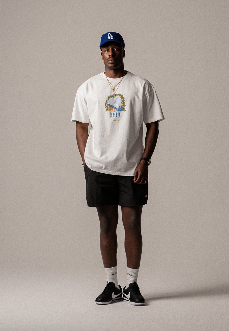 T-shirt en coton blanc avec un design graphique coloré, shorts noirs et baskets noires avec des accents blancs. Casquette bleue et chaussettes blanches arrivant aux chevilles.