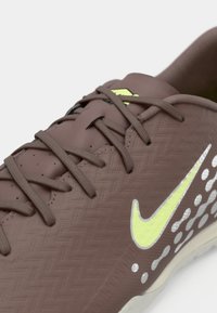 Καφέ αθλητικό παπούτσι με υφή υλικού, έντονο κίτρινο λογότυπο Nike, γκρι λεπτομέρειες και κλείσιμο με κορδόνια. Σχεδιαστικό μοτίβο στο πλάι.
