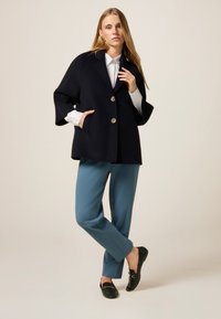 Cappotto a mantello in misto lana blu navy con due bottoni dorati, abbinato a pantaloni teal e mocassini neri con superficie testurizzata.