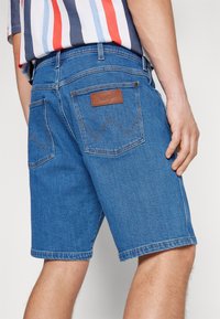 Modré džínové šortky se rovnýcm střihem, které mají dvě zadní kapsy, koženou nášivku "Wrangler" a kontrastní šití v klasickém designu.