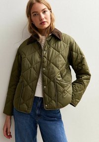 Jonge vrouw in een olijfgroene gewatteerde jas met een bruine kraag, witte trui en blauwe spijkerbroek, poseert tegen een lichte achtergrond.