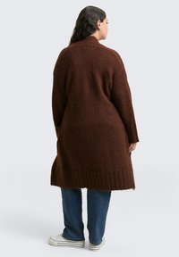 Brązowy sweter na drutach o fakturowanym wykończeniu, z długimi rękawami, ściągaczem na dole i luźnym krojem, zestawiony z niebieskimi dżinsami i białymi trampkami.