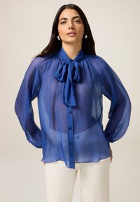 Durchsichtiges blaues Blusenshirt mit einem Krawattenkragen, Knopfleiste und langen Ärmeln. Glatte Textur mit leichtem und durchscheinendem Aussehen.