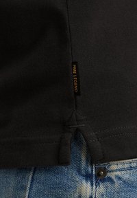 Zwarte katoenen T-shirt met een label "PME Legend" aan de naad, gepresenteerd boven lichtblauwe denim met een zichtbare knoop.