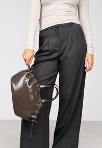 Bolso de mano marrón y brillante hecho de cuero con forma redondeada, detalles de cordones y asas dobles, sostenido por una persona que lleva pantalones grises.