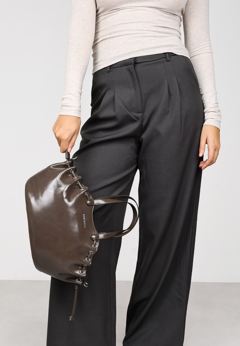 Bolso de mano marrón y brillante hecho de cuero con forma redondeada, detalles de cordones y asas dobles, sostenido por una persona que lleva pantalones grises.