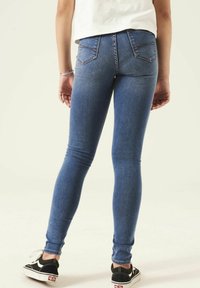 Jeans ajustados en denim azul, con un diseño de tiro medio, cinco bolsillos, costuras en contraste y un ligero desgaste. Combinados con zapatillas negras.