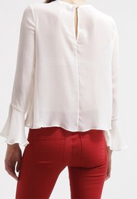 Blusa bianca a pieghe con maniche a campana e dettaglio a apertura a chiave sul retro, indossata sopra pantaloni rossi attillati. Tessuto liscio e materiale leggero.