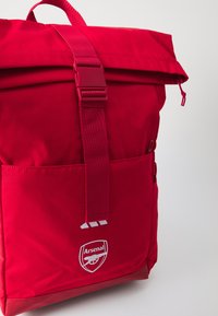 Red backpack with a roll-top design, adjustable strap, front zip pocket, and Arsenal logo. Made of durable fabric with a smooth texture.