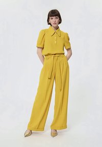 Donna che indossa una blusa a maniche corte giallo senape con colletto ampio e pantaloni larghi a vita alta abbinati con cintura, in piedi con le mani dietro la schiena.