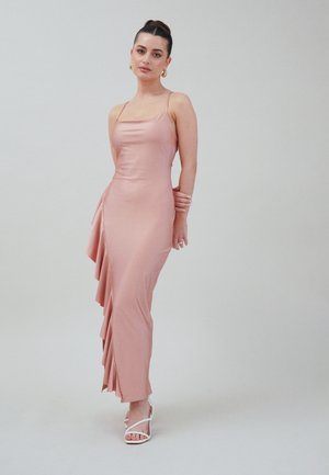 Rochie de satin roz deschis, cu un corset ajustat, bretele subțiri și volane drapate asimetrice pe o parte; asortată cu sandale albe cu curea.