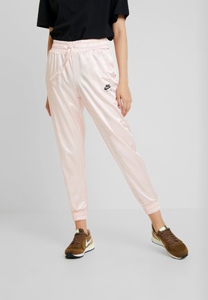Pantalons de jogging en satin rose clair avec taille et poignets élastiques, arborant un petit logo Nike noir sur le côté gauche. Portés avec des baskets marron.