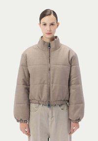 Vadderad jacka i ljusbrunt tyg med hög krage, med en framdragkedja samt elastiska manschetter och nederkant. Bärs över ljusa byxor.