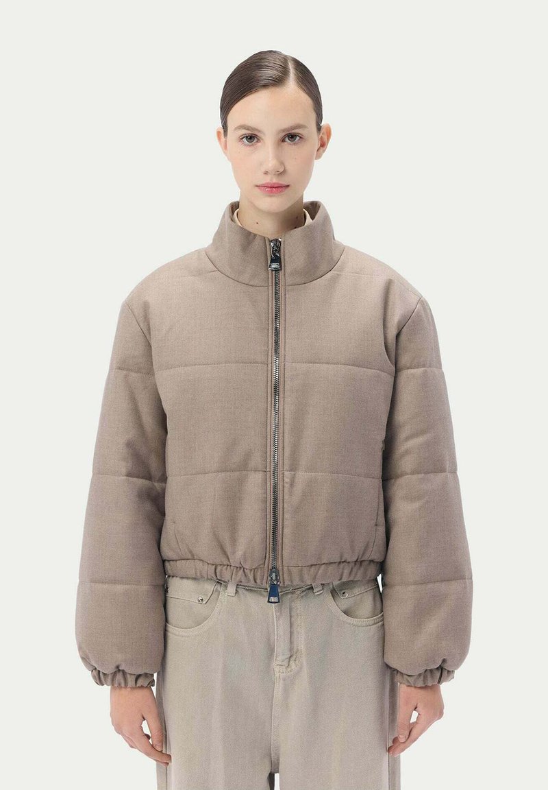 Vadderad jacka i ljusbrunt tyg med hög krage, med en framdragkedja samt elastiska manschetter och nederkant. Bärs över ljusa byxor.