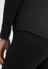 Zwarte longsleeve shirt met ribbelstructuur, gebogen zoom en labeldetail, gecombineerd met zwarte leggings. Close-up van de onderzijde.