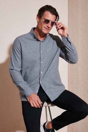 Camisa gris de manga larga con botones y cuello, combinada con pantalones negros. El modelo lleva gafas de sol y está sentado en un taburete.