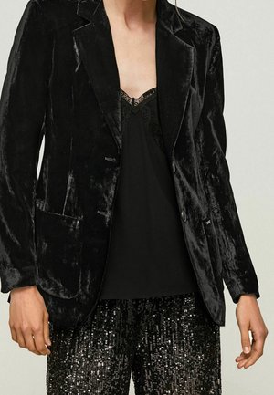 Frau trägt schwarzen Samtblazer über schwarzem Spitzen-Top mit Spaghettiträgern und schwarze Paillettenhose, steht vor neutralem Hintergrund.