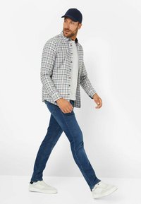 Karierte Hemdbluse in Blau, Weiß und Braun, über einem weißen T-Shirt getragen. Dunkelblaue Jeans und weiße Sneaker runden das Outfit ab.