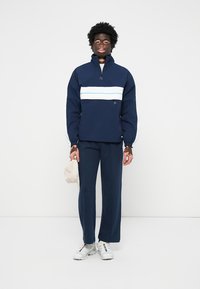 Marineblå pulloverjakke med en hvid vandret stribe, parret med matchende marineblå bukser og hvide sneakers. Let stof, afslappet pasform, elastiske detaljer.