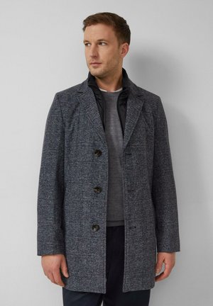 Mann trägt einen dunkelgrauen, karierten Mantel mit schwarzem, gestepptem Innenkragen über einem grauen Pullover, steht vor einem einfarbigen grauen Hintergrund.