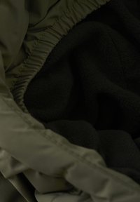 Un tessuto verde con una superficie liscia si fonde in un morbido interno in pile scuro. Presenta una coulisse elastica lungo il bordo.
