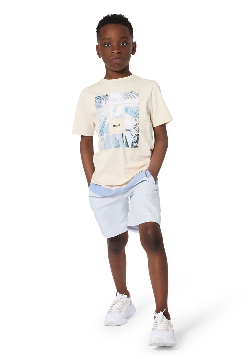 T-shirt beige avec graphique bleu et logo "BOSS", associé à un short bleu clair et des baskets blanches, avec poches et coupe décontractée.