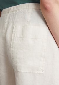 Pantalones cortos de lino con cintura elástica, que cuentan con un bolsillo parche en la parte trasera. El color es blanco roto con una textura suave.