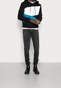 Černá mikina s bílým a tyrkysovým geometrickým vzorem, spojená se štíhlými tmavě šedými džínami a černými kotníkovými botami. Hladká textilní struktura.