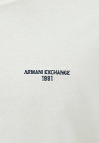 Baltas medvilninis marškinėliai su "ARMANI EXCHANGE 1991" siuvinėjimu tamsiai mėlyna spalva kairėje pusėje. Lygus tekstūras, minimalistinis dizainas.