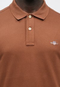 Bruin poloshirt van gestructureerde stof met een puntige kraag, voorzien van twee knopen en een klein GANT-logo geborduurd op de borst.