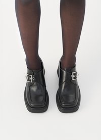 Pantofi din piele neagră cu vârf pătrat, talpă groasă și un accent cu cataramă argintie ajustabilă. Purtați cu ciorapi negri subțiri.