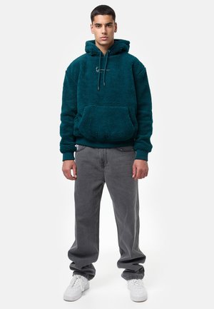 Türkiser Fleece-Hoodie mit einer vorderen Kängurutasche, Kapuze mit Kordelzug und gerippten Bündchen, kombiniert mit grauen, locker sitzenden Jeans und weißen Sneakers.