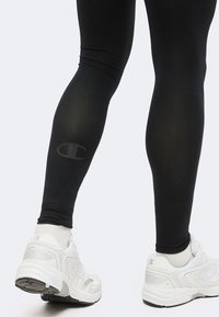 Jambes portant des leggings noirs avec un discret logo Champion sur le mollet, assortis à des baskets blanches Champion et des chaussettes blanches sur un fond uni.