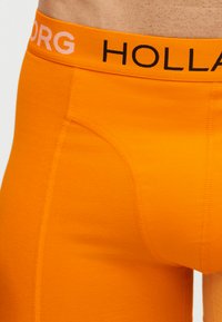 Κοντινό πλάνο ενός ατόμου που φορά φωτεινό πορτοκαλί μπόξερ με ζώνη μέσης όπου αναγράφεται «HOLLAND» με μαύρα και λευκά γράμματα.