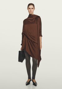 Bruine asymmetrische jurk met gedrapeerde stof, driekwart mouwen, gecombineerd met donkergrijze legging en zwarte handtas. Platte zwarte schoenen.