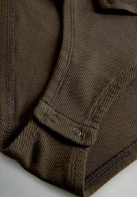 Ruda gofruota medžiaga su sagos užsegimo detalėmis ir iškilusiu siuvimu. Medžiagos tekstūra yra minkšta, o užfiksuotas artimas vaizdas išryškina raštą.