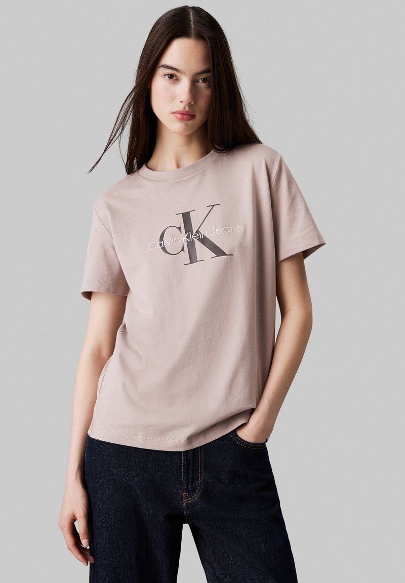 Light pink cotton T-shirt featuring a large 'CK' logo and "Calvin Klein Jeans" text. Model wears dark jeans, standing against a neutral background.
