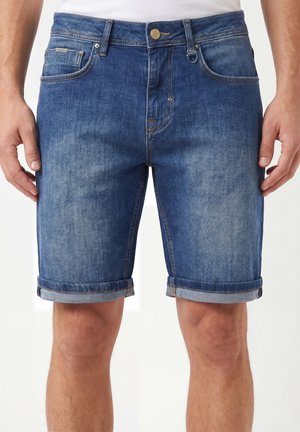 Hombre con pantalones cortos de mezclilla azul medio con dobladillos enrollados y costuras visibles, de pie con las manos a los lados contra un fondo liso.