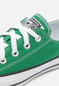 Adidași verzi din pânză cu șireturi albe, oglinzi argintii și o dungă neagră pe talpă. Include un patch logo proeminent pe limbă.
